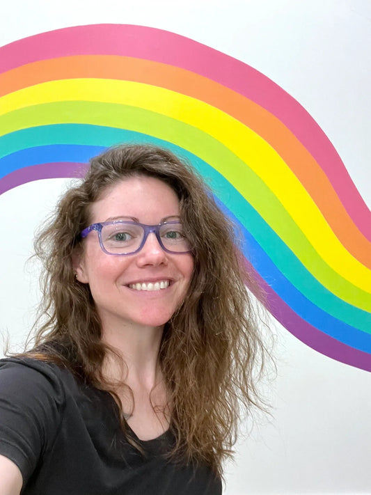 Camille Gerrick smiling, wearing purple glasses, standing in front of rainbow wall mural