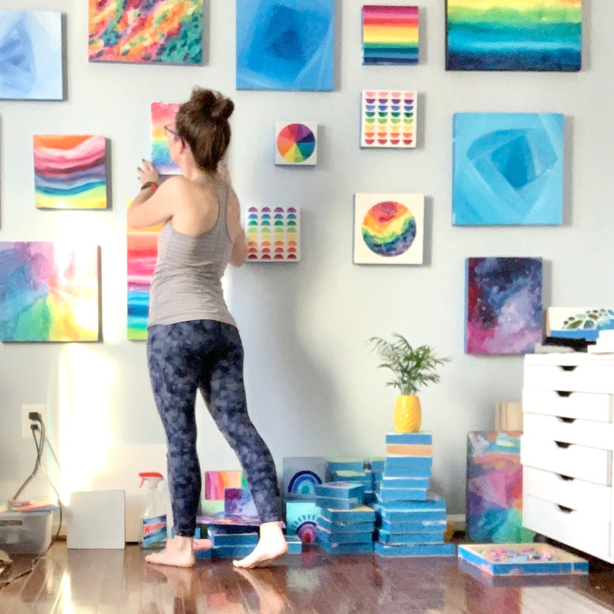 Camille hanging up another one of her colorful paintings on a gallery wall in her studio. Paintings include rainbow gradients and colorwheels