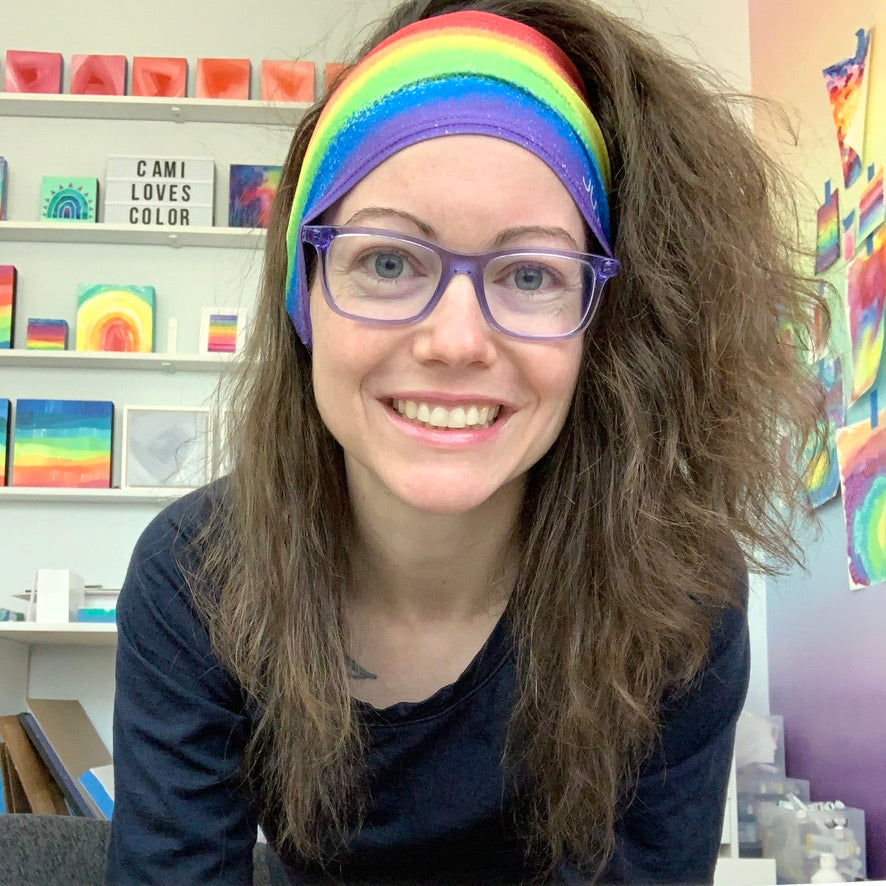Camille wearing a rainbow head band in her studio, smiling, with her colorful art behind her