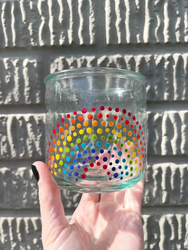 Hand holding a clear glass cup with hand-painted dotted ROYGBIV rainbow by Camille Gerrick in daylight in front of concrete wall