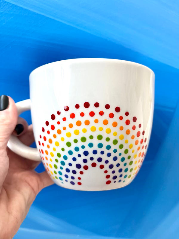 Hand holding a Porcelain Colorful Short Latte Mug with hand-painted dotted ROYGBIV rainbow by Camille Gerrick with blue background