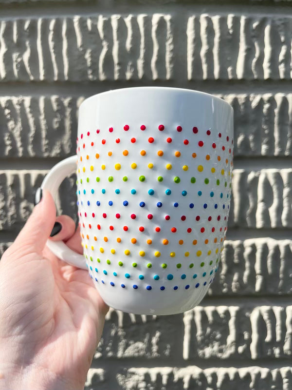Person holding Porcelain Vibrant Rainbow Dot Tall Latte Mug with bands of rainbow dots wrapping around it. Made by Camille Gerrick. In direct sunlight, in front of concrete wall