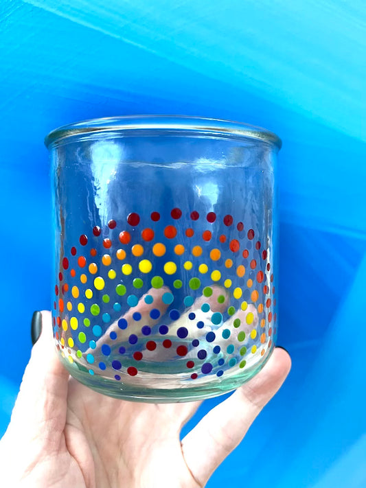 Hand holding a clear glass cup with hand-painted dotted ROYGBIV rainbow by Camille Gerrick in front of blue background