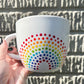 Hand holding a Porcelain Colorful Short Latte Mug with hand-painted dotted ROYGBIV rainbow by Camille Gerrick in direct sunlight in front of concrete wall