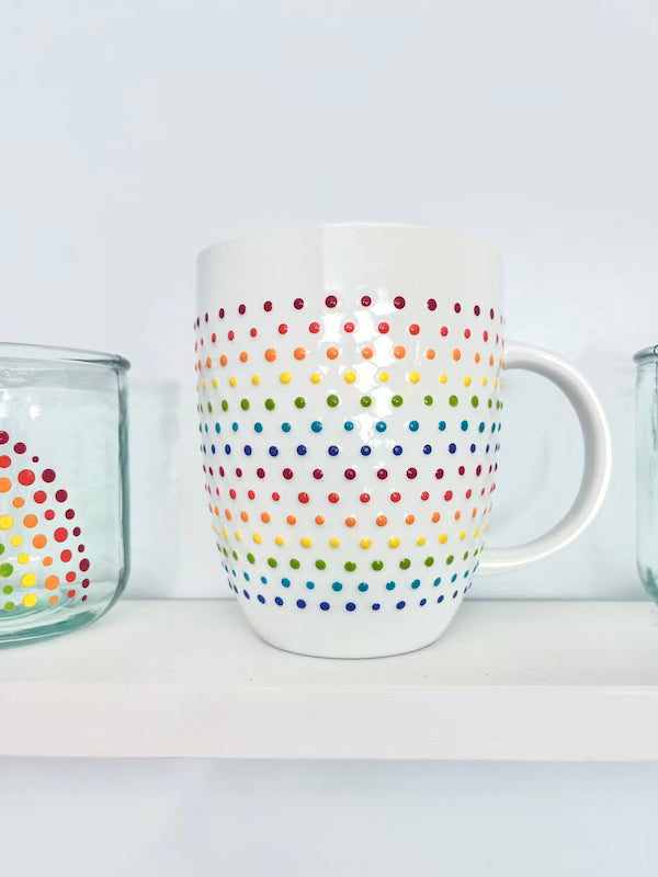 Porcelain Vibrant Rainbow Dot Tall Latte Mug with bands of textured rainbow dots wrapping around it on a white shelf. Made by Camille Gerrick.