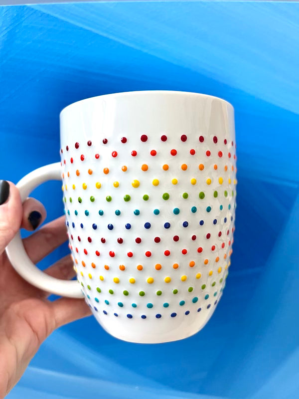 Person holding Porcelain Vibrant Rainbow Dot Tall Latte Mug with bands of rainbow dots wrapping around it. Made by Camille Gerrick. In front of blue background